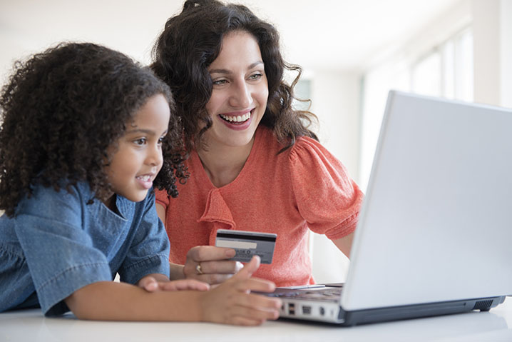 Mulher com um cartao na mao olhando para um computador com sua filha